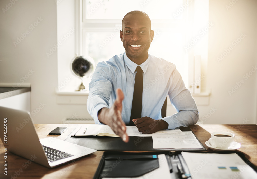 Smiling young executive sitting at his desk extending a handshak