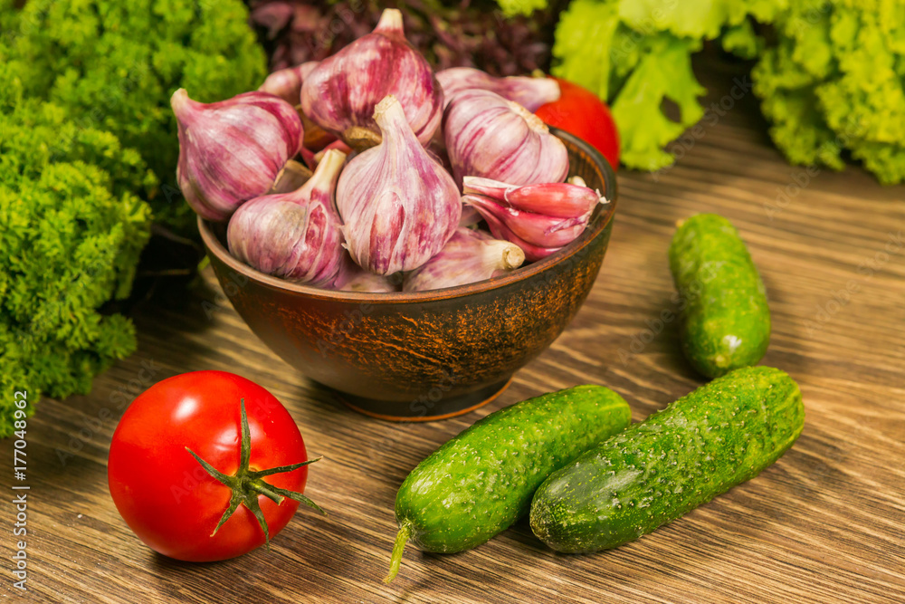 A full bowl with garlic bulbs. Tomatoes and cucumbers on a wooden table. Green salad in the backgrou