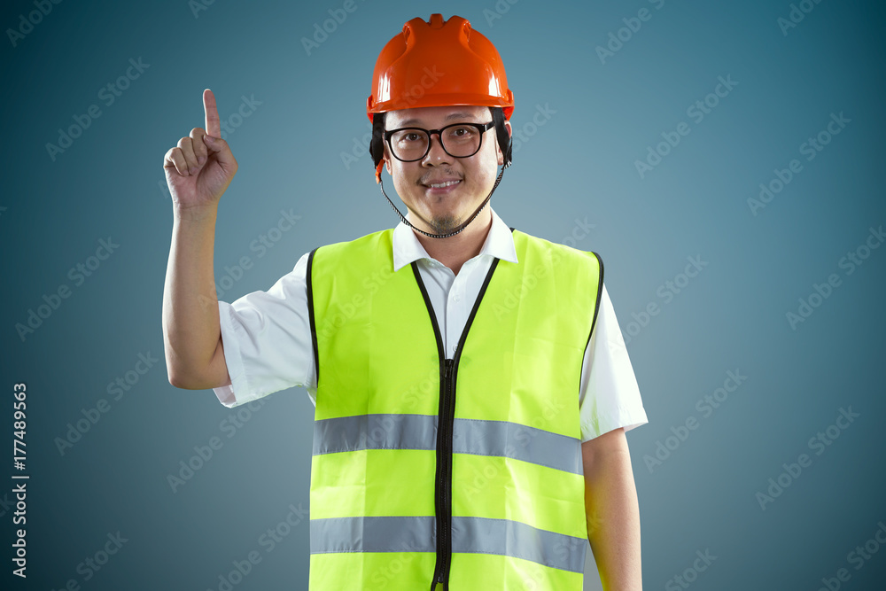 Young asian construction workers in safety helmet pointing up the finger ,isolated on blue backgroun