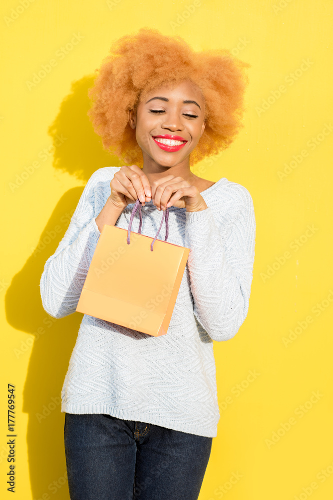 Portrait of a beautiful african woman in blue sweater standing with shopping bag on the yellow backg