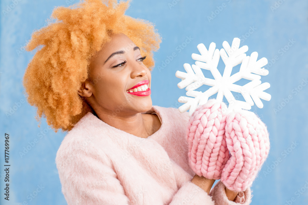 Portrait of a cute african woman in the pink coat holding an artificial snowflake on the blue wall b