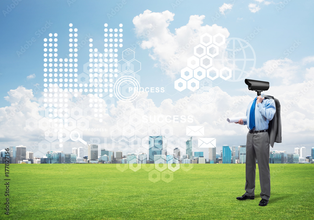 Camera headed man standing on green grass against modern cityscape