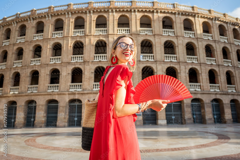 Woman in red dress with spanish hand fan stnading back in front of the bullring amphitheatre in Vale