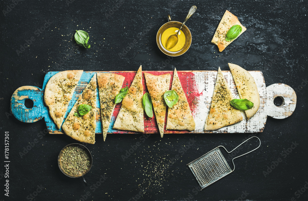 Homemade Italian focaccia flatbread cut into pieces with herbs, fresh basil leaves, olive oil and gl