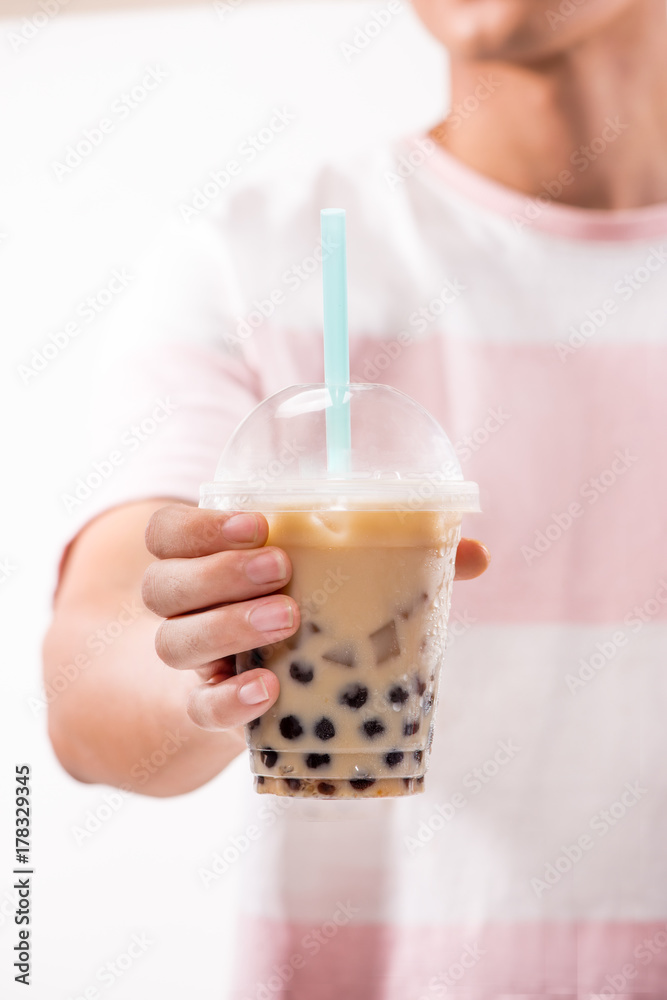 Hand holding light brown creamy bubble tea and black tapioca pearls in plastic cups on table.