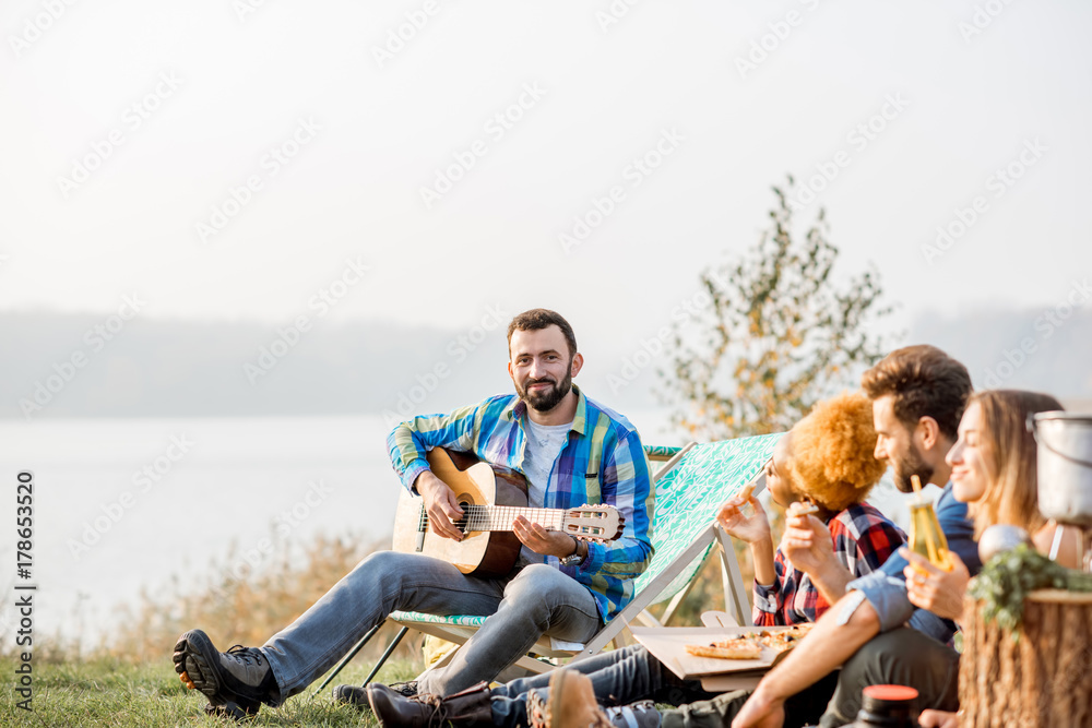 Multi ethnic group of friends dressed casually having a picnic, playing guitar and eating pizza, dur