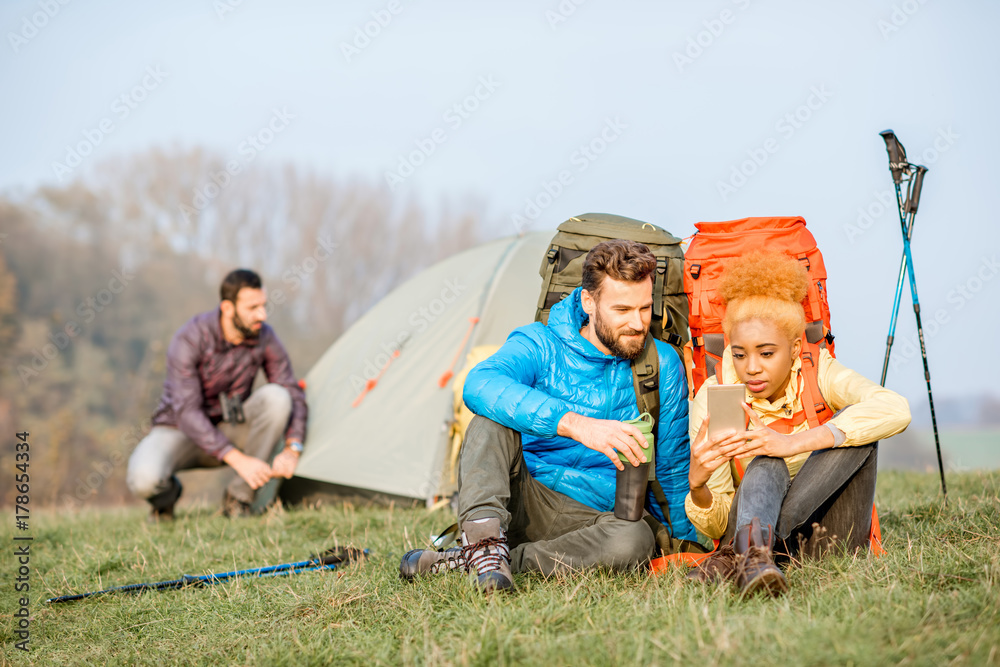 年轻夫妇穿着五颜六色的夹克，背着背包，坐在绿色法律上玩智能手机