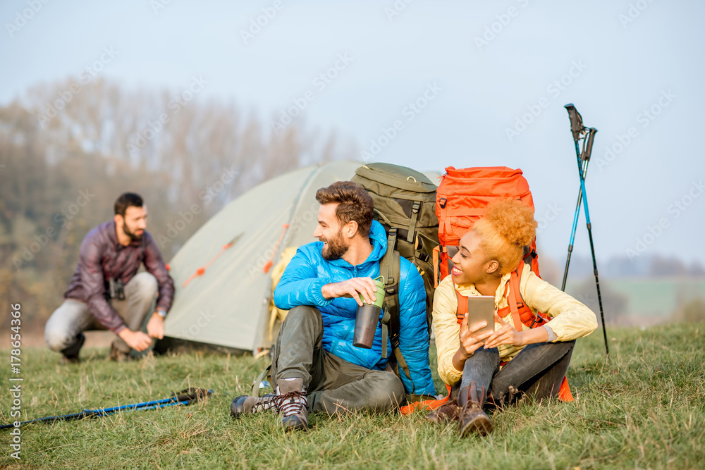 年轻夫妇穿着五颜六色的夹克，背着背包，坐在绿色法律上玩智能手机