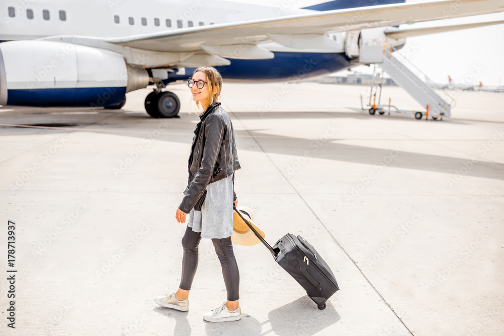 Young woman in leather jacket walking with suitcase near the airplane outdoors on the airport runway