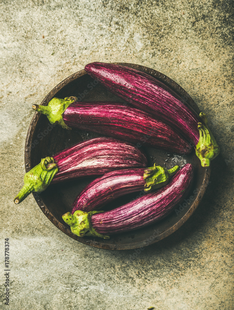 Flat-lay of fresh raw Fall harvest purple eggplants or aubergines in wooden bowl over concrete stone