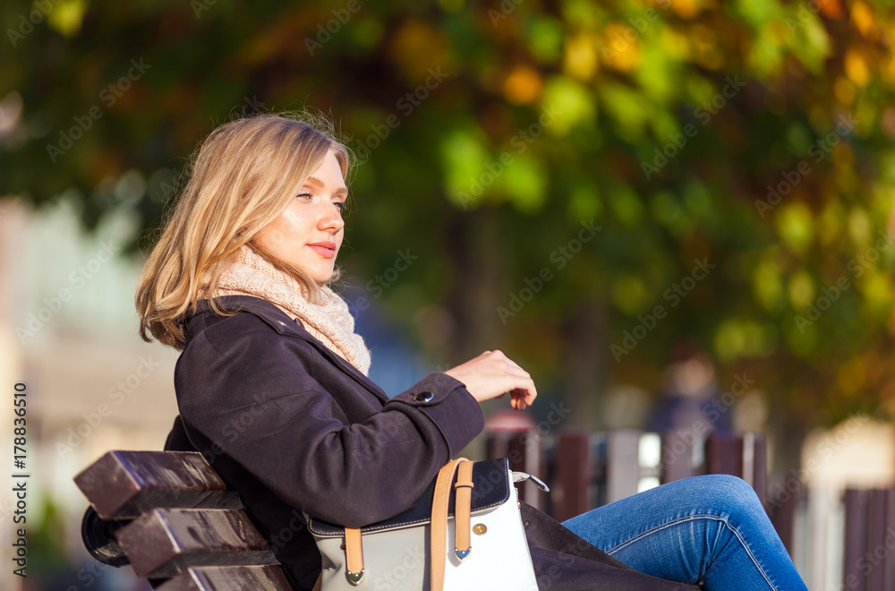 Young woman sitting on bench at the city street during sunny autumn day