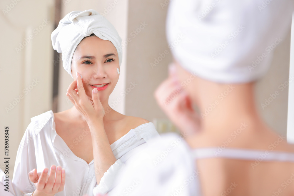 Young asia woman applying foundation or moisturizer on her face in front of mirror.