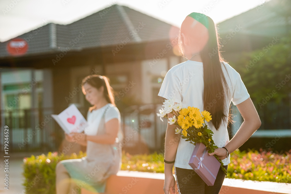 The girl, which is the daughter, flower and gift box is hidden behind her to bring it to her mother 