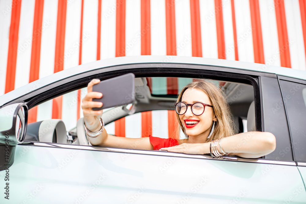 Young and happy woman making selfie photo with smart phone sitting at the car on the red wall backgr