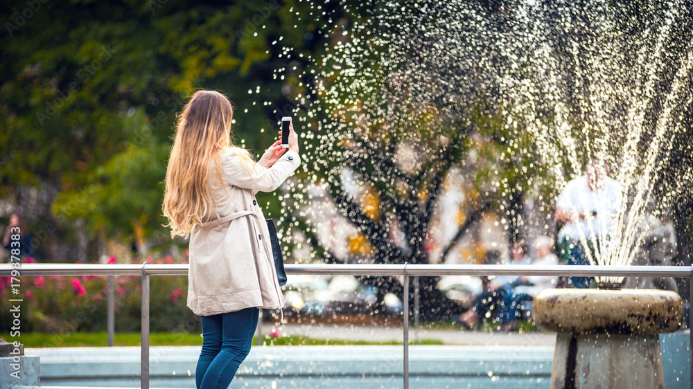 Woman in the autumn city taking picture of fountain on smartphone