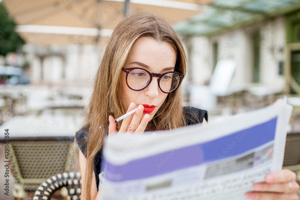 Close-up portrait of a young and elegant woman smoking a cigarette sitting outdoors with newspaper a