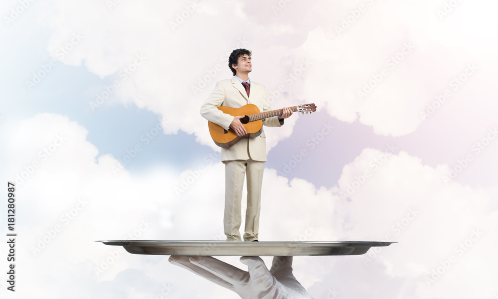 Businessman on metal tray playing acoustic guitar against blue sky background