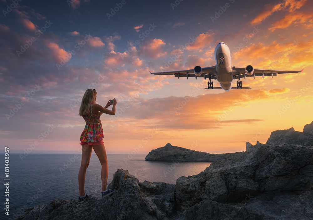 Beautiful woman makes photo of landing aircraft at sunset. Summer landscape with girl standing on th