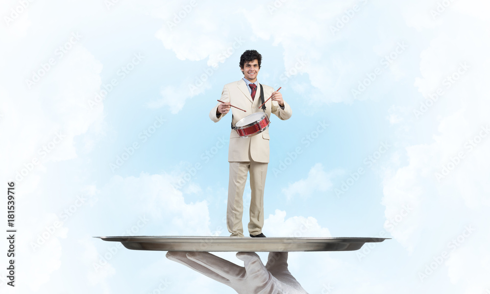 Young businessman on metal tray playing drums against blue sky background