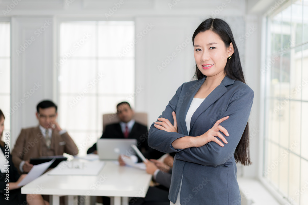 Happy businesswoman standing with arms crossed at office while colleagues discussing in background.