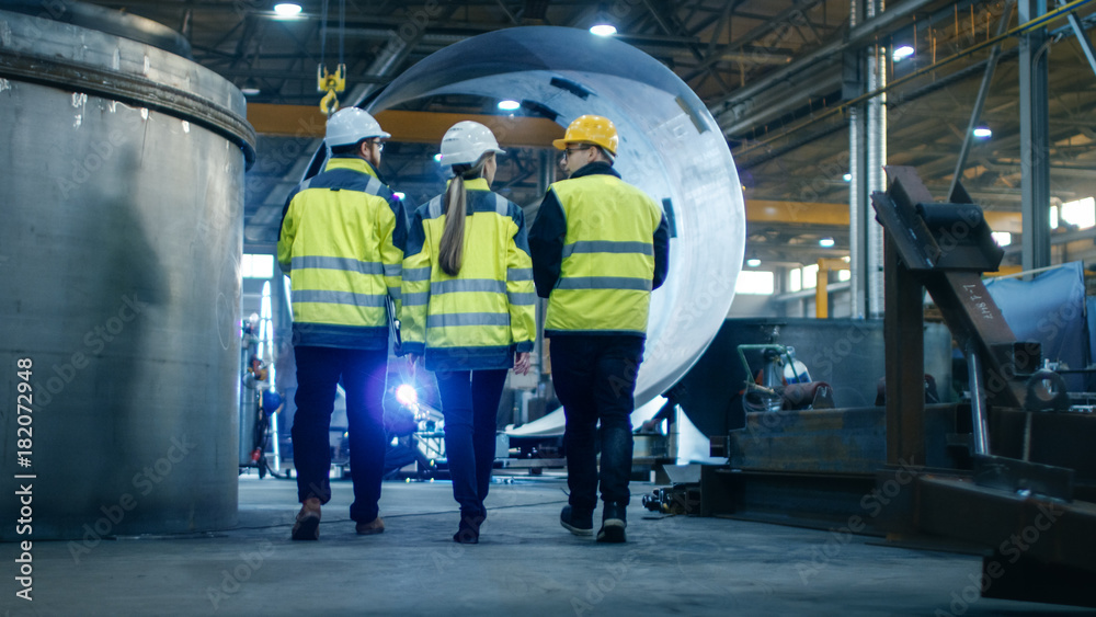 Following Shot of Three Engineers Walking Through Heavy Industry Manufacturing Factory. In the Backg