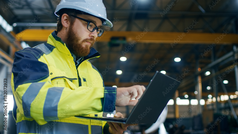 Industrial Engineer in Hard Hat Wearing Safety Jacket Uses Touchscreen Laptop. He Works at the Heavy