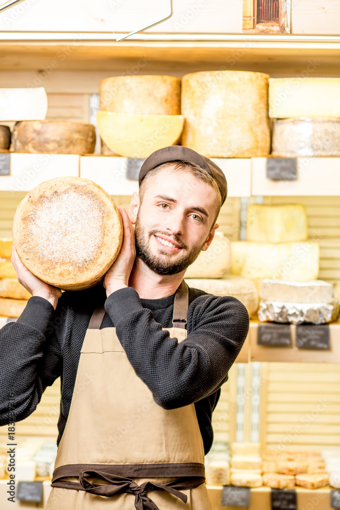 Portrait of a handsome cheese seller in uniform holding a big seasoned cheese in front of the store 