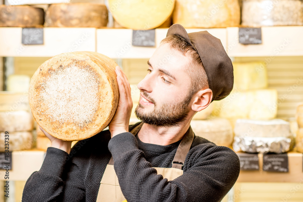 Portrait of a handsome cheese seller in uniform holding a big seasoned cheese in front of the store 