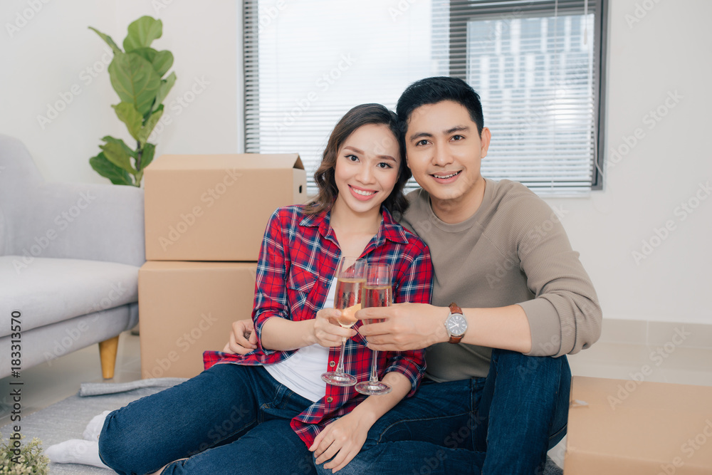 Couple lovers moving into new home sitting among cardboard boxes enjoying wine