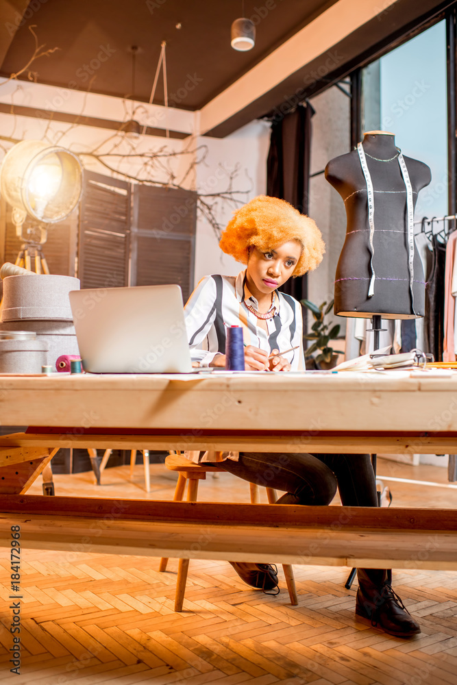 Young african fashion designer working with clothes drawings and laptop sitting at the studio interi