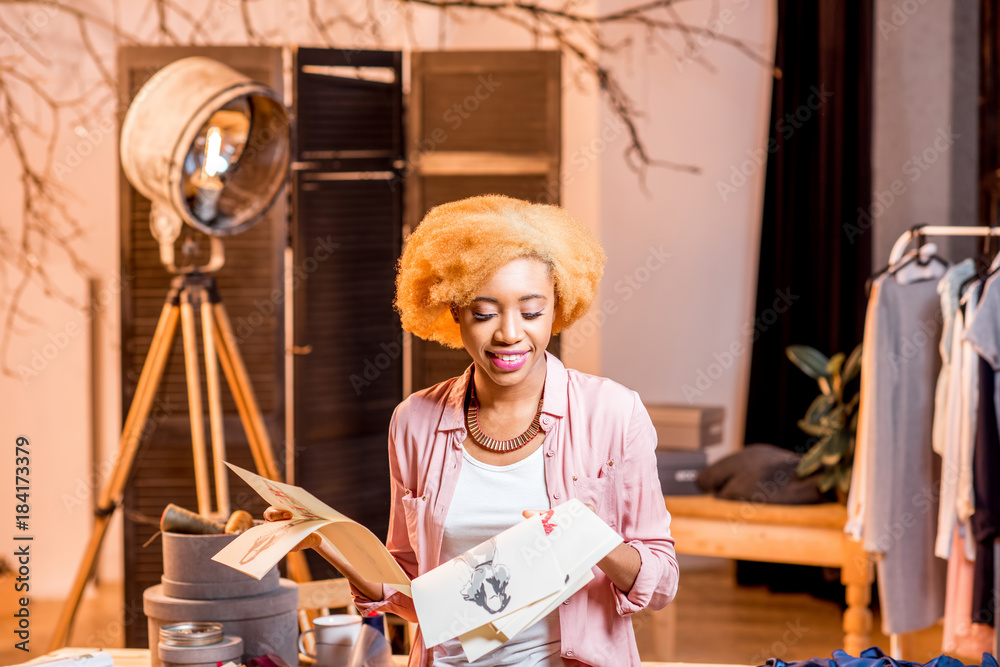 Young african fashion designer working with clothes drawings and laptop sitting at the studio interi