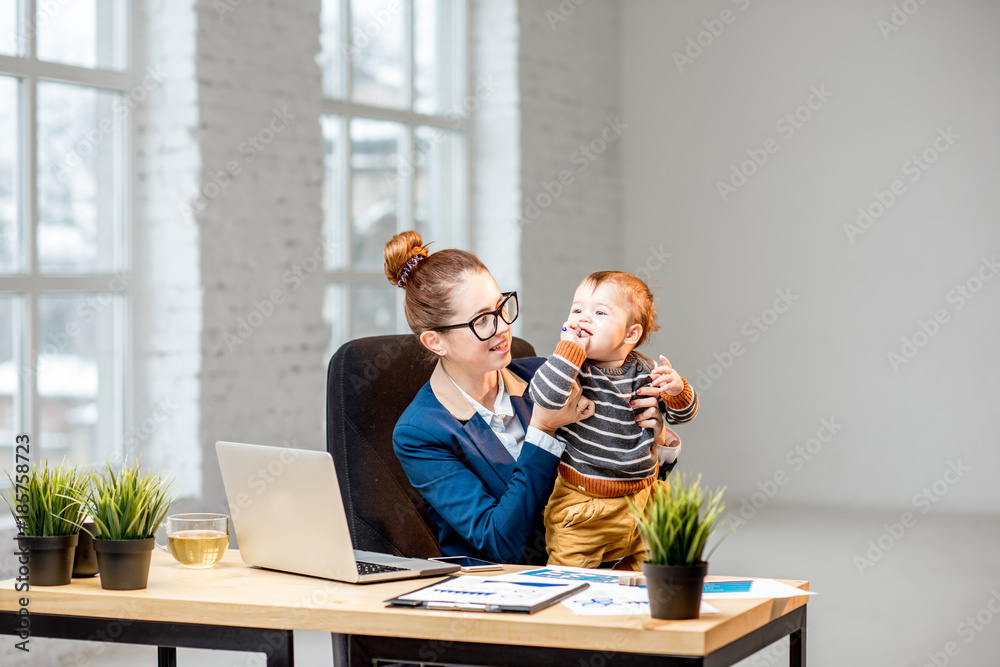 Young multitasking businessmam dressed in the suit having fun with her baby son at the office