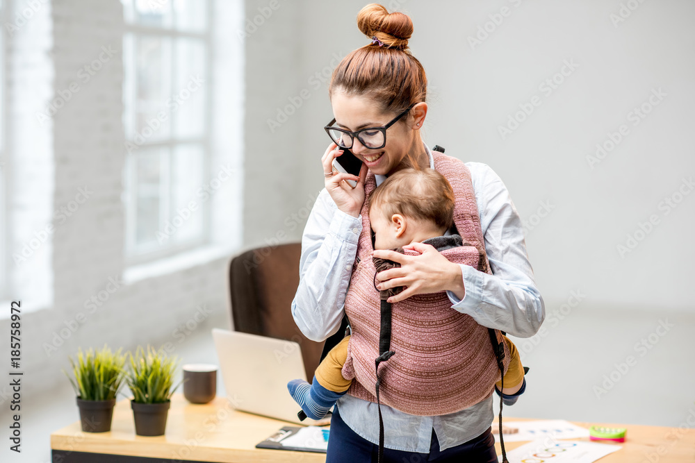 年轻的多任务女商人在办公室工作时带着她的小儿子站着