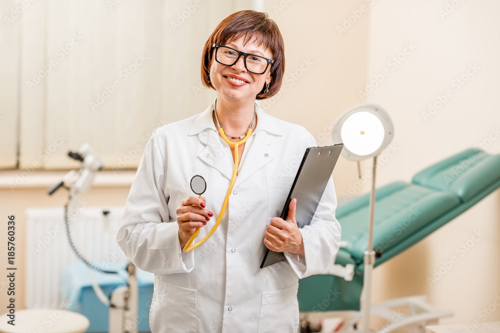 Portrait of a senior woman gynecologist standing in the office with gynecological chair and lamp on 