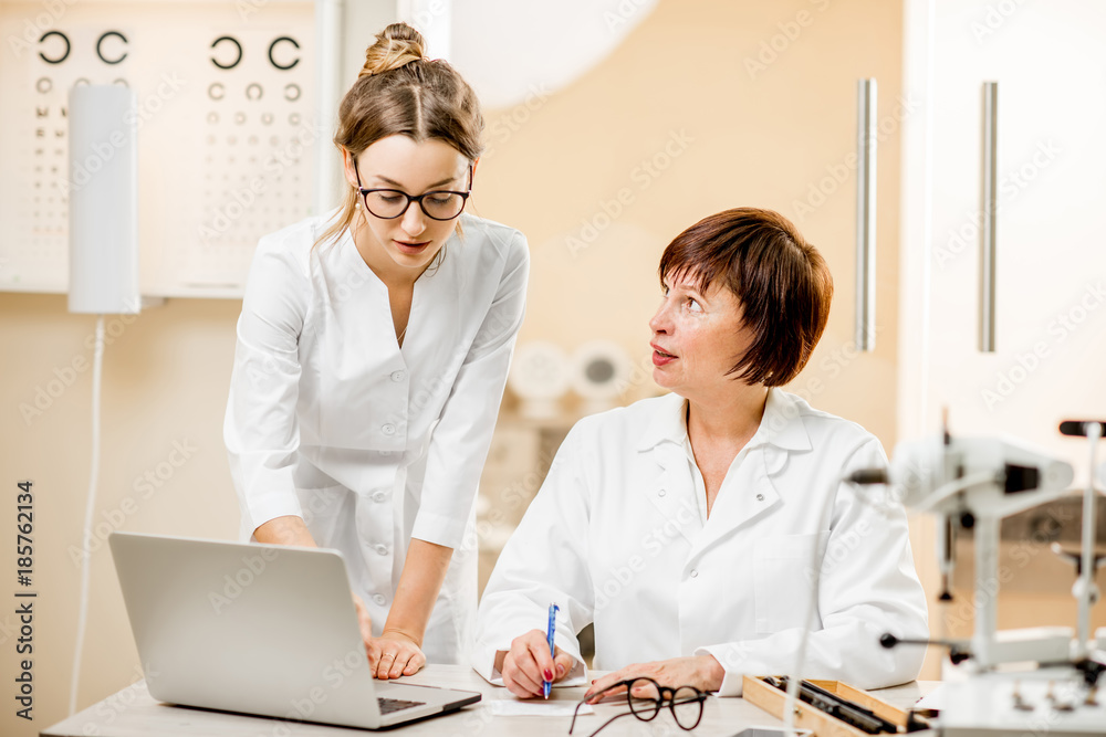 Senior woman ophthalmologist with young female assistant working with laptop at the office