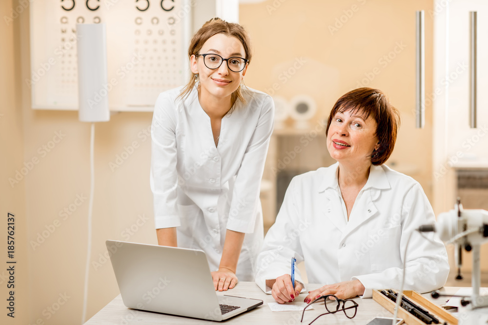 Senior woman ophthalmologist with young female assistant working with laptop at the office