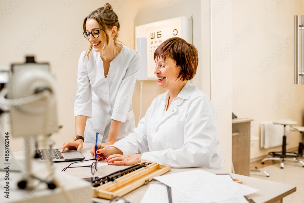 Senior woman ophthalmologist with young female assistant working with laptop at the office