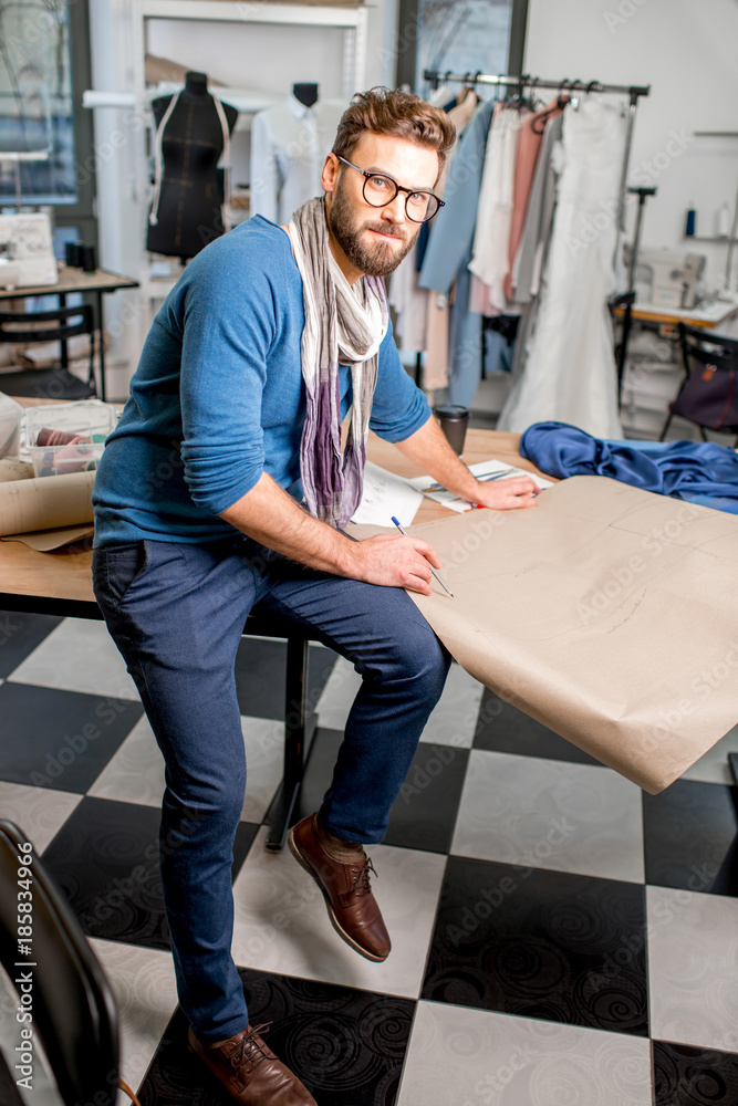 Portrait of a handsome fashion designer sitting with paper sketches at the studio full of tailoring 