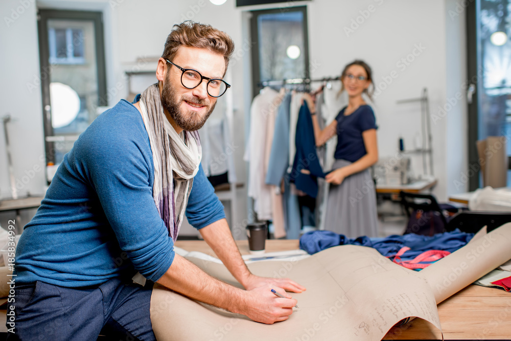 Portrait of a handsome fashion designer sitting with paper sketches at the studio full of tailoring 