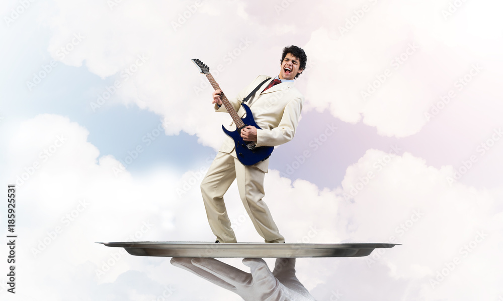 Businessman on metal tray playing electric guitar against blue sky background