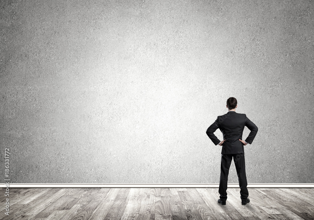 Businessman with hands on waist standing with back and looking at blank concrete wall