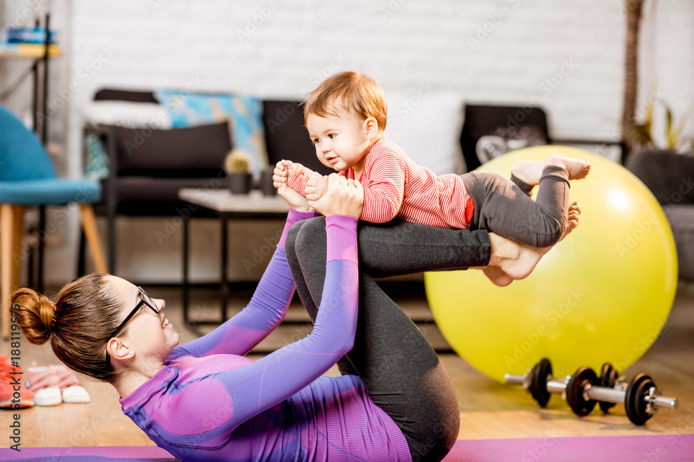 Young mother in sportswear doing exercise lifting with legs her baby son on the mat at home