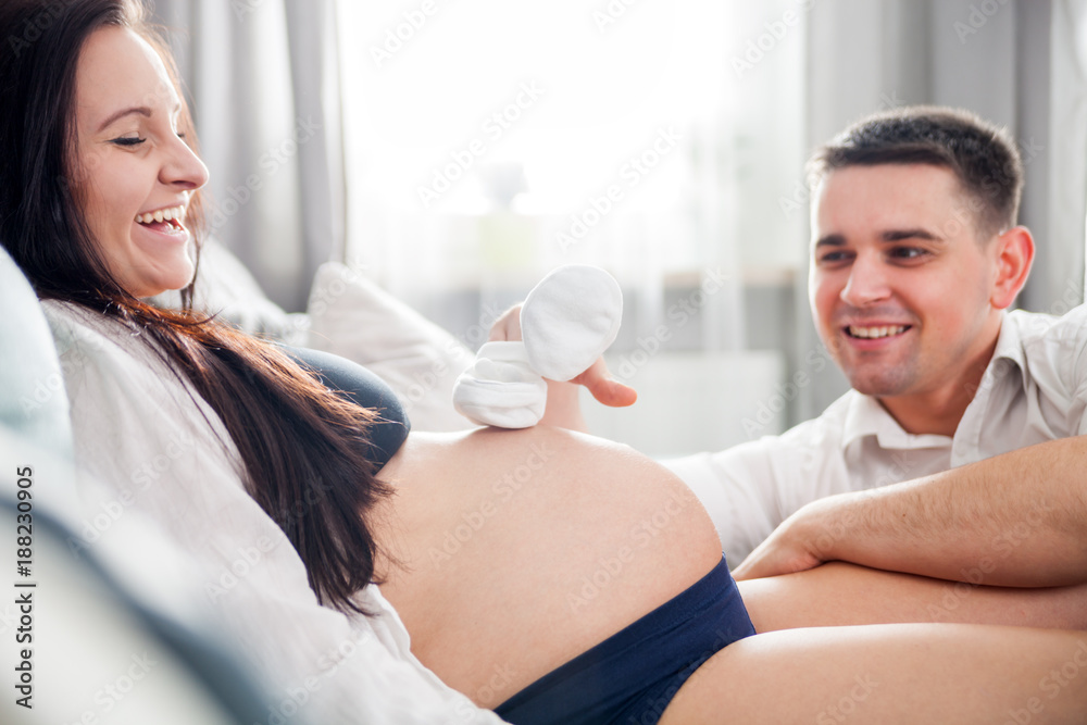 Pregnant couple playing together with baby shoes sitting on sofa at home