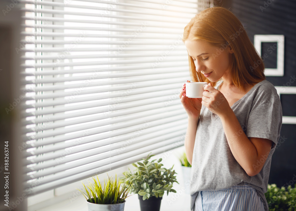 Happy young woman meets   morning with  cup of coffee  at   window