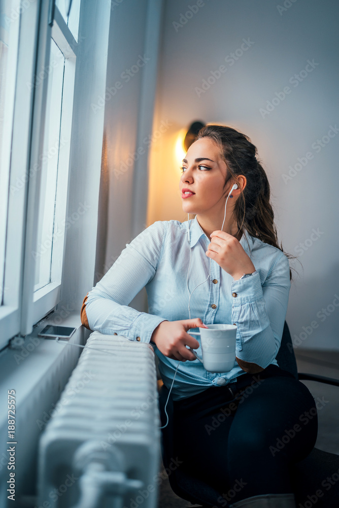 Cheerful young girl listening to music with headphone near window and holding cup of coffee.