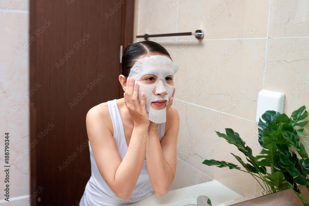 Asian woman applying mask on her face and looking in the mirror in the bathroom