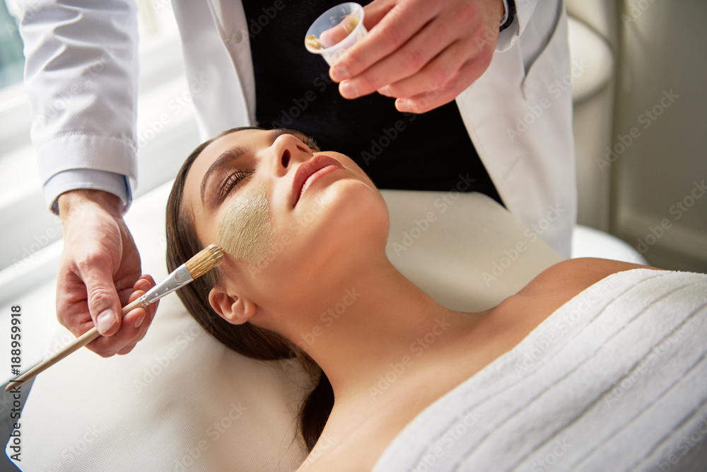 Woman lying with eyes closed while beautician putting mask