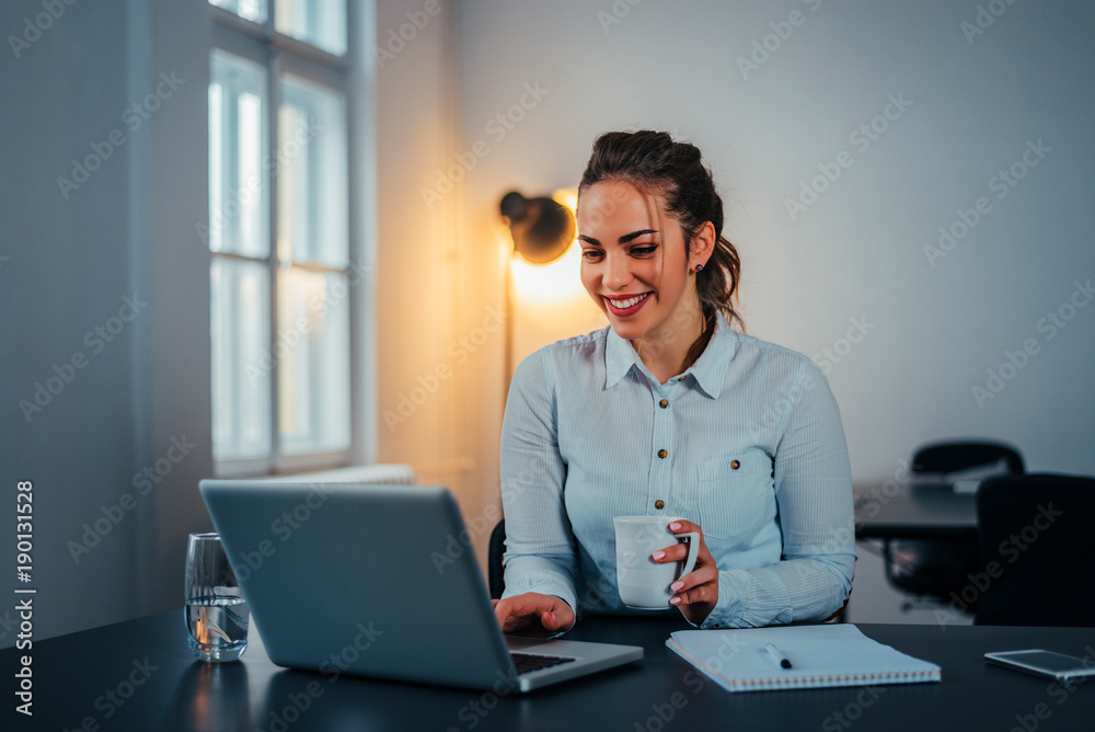 年轻的女商人坐在桌子上，手里拿着杯子和笔记本电脑。