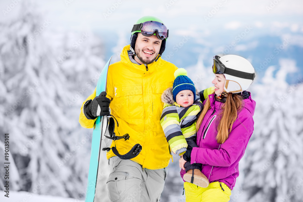 Portrait of a happy family with baby boy in winter sports clothes standing with snowboard during the