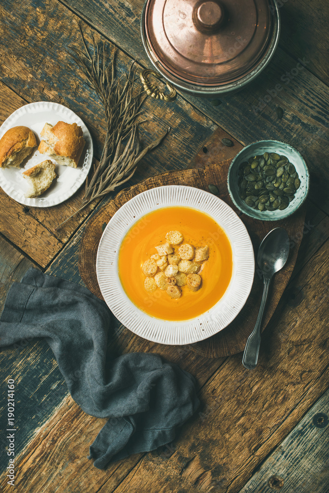 Flat-lay of fall warming pumpkin cream soup with croutons and seeds on board over rustic wooden back